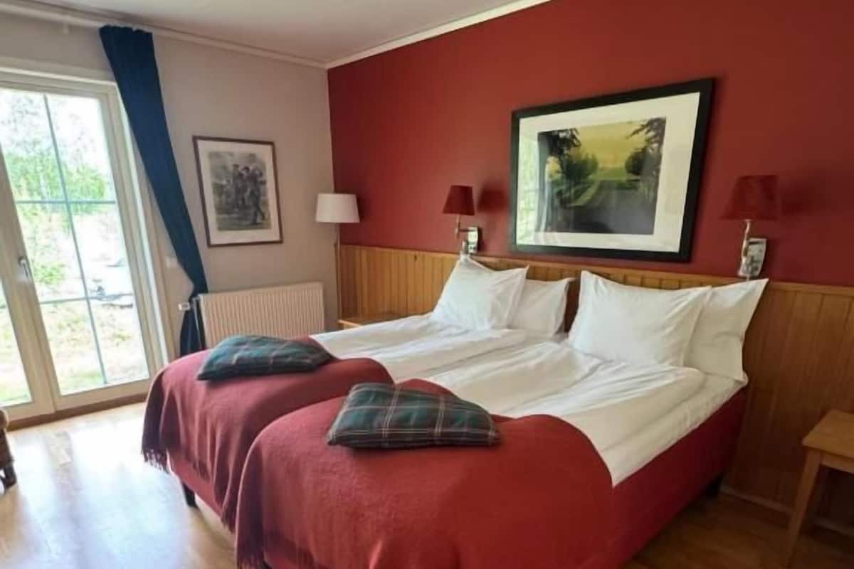 A double bed with red bedding and two pillows, a wooden headboard, a painting on the wall, a window with curtains, and a radiator.