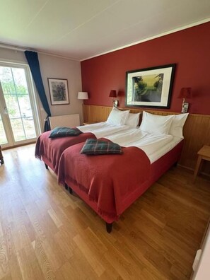 A double bed with red bedding and two pillows, a wooden headboard, a painting on the wall, a window with curtains, and a radiator. - Ada Krog Hotell & Konferens (Trosa)