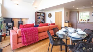 A modern living room with a sofa, a dining table set for four, and a kitchen area with a stove. - Howells Mere 16, Indian Summer - Lakeside Cottage | Sleeps 4 in 2 bedrooms (Somerford Keynes)