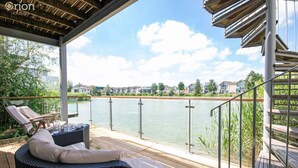 A covered outdoor deck with wicker furniture and a view of a lake and residential buildings. - Howells Mere 16, Indian Summer - Lakeside Cottage | Sleeps 4 in 2 bedrooms (Somerford Keynes)