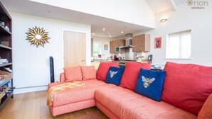 A cozy living room with a sofa, a decorative wall piece, and a view into the kitchen. - Howells Mere 16, Indian Summer - Lakeside Cottage | Sleeps 4 in 2 bedrooms (Somerford Keynes)