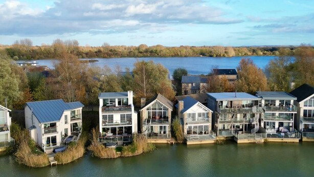 A lakeside residential area with modern houses featuring balconies and large windows, surrounded by trees and a calm lake. - Howells Mere 16, Indian Summer - Lakeside Cottage | Sleeps 4 in 2 bedrooms (Somerford Keynes)