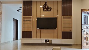 A modern living room with a flat-screen TV mounted on a wooden wall, surrounded by built-in shelves and a glass coffee table. - Pawna Ns4 villa (Mawal)