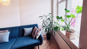 A blue sofa with patterned cushions, a potted plant, and a window with a view of buildings. - Nordic Vintage Studio (Helsinki)