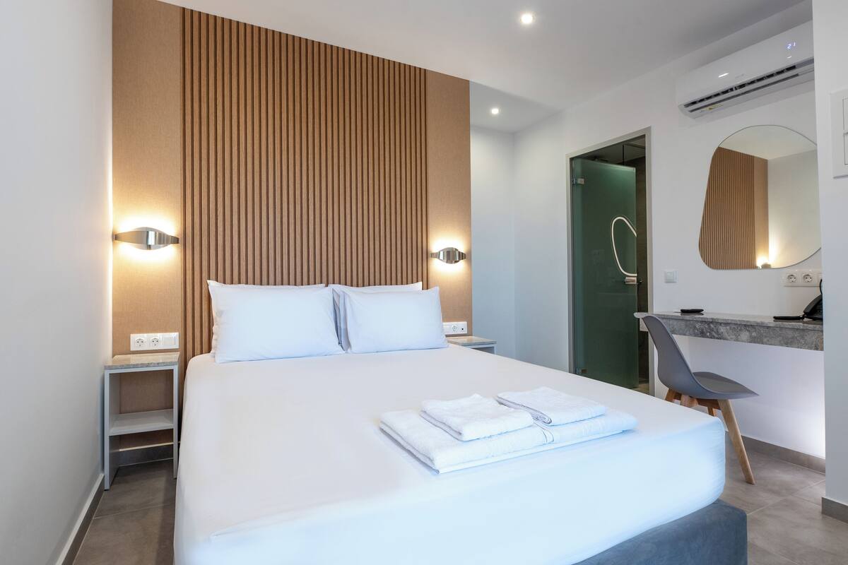 A modern hotel room with a large bed, a bedside table, a nightstand, a bathroom with a mirror, and a small seating area.