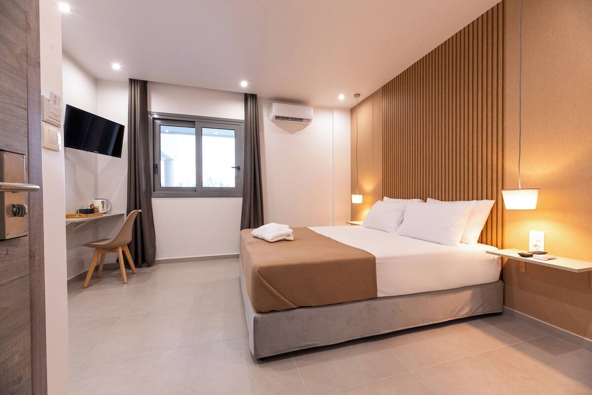 A modern hotel room with a bed, bedside table, lamp, TV, and a desk with chair.