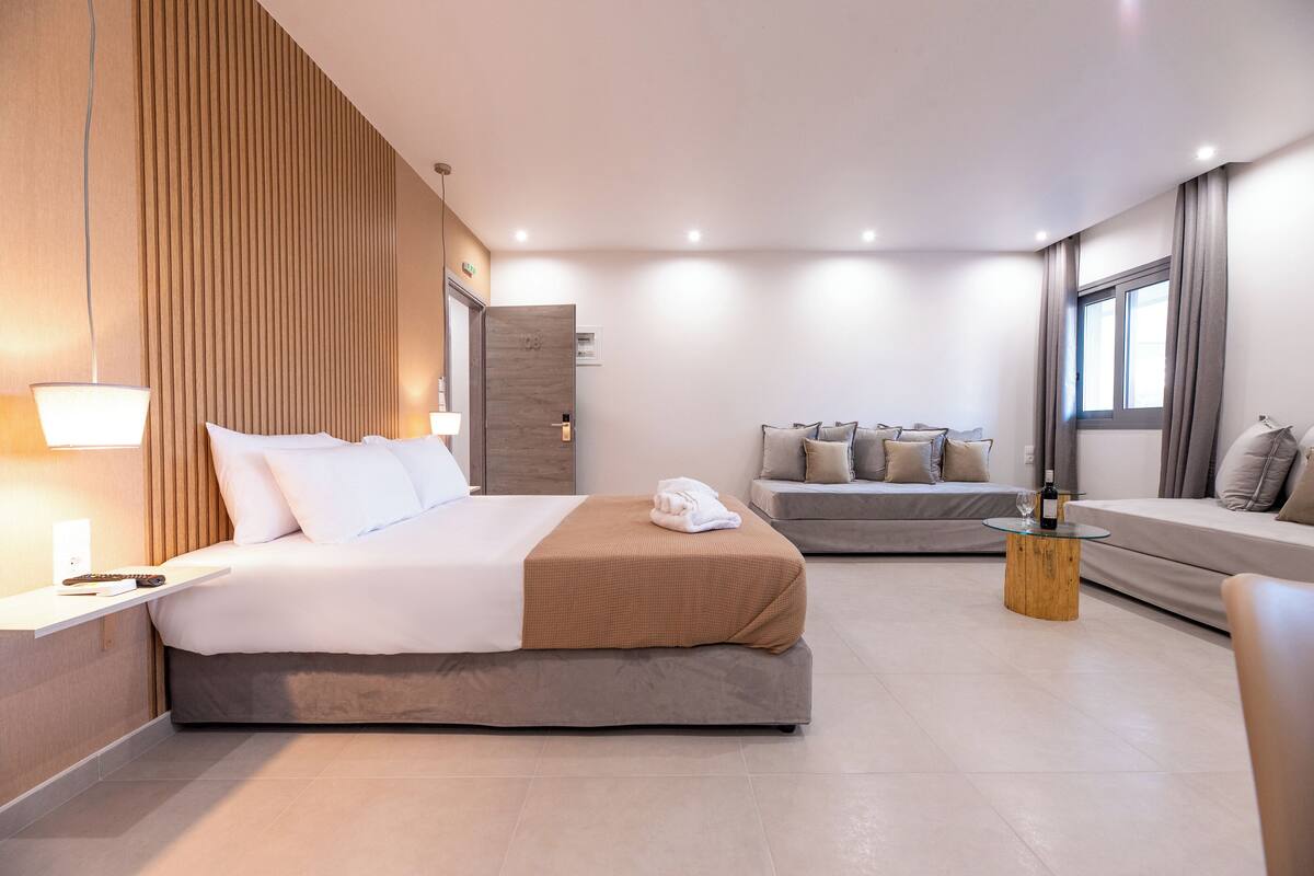 A modern hotel room with a large bed, a smaller sofa, a wooden coffee table, and a view of the city.