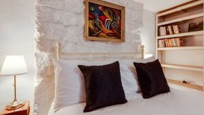 A bedroom with a white stone wall, a bed with white and dark pillows, a painting, and a bookshelf. - Basement Studio in Summertown with kitchenette (Oxford)