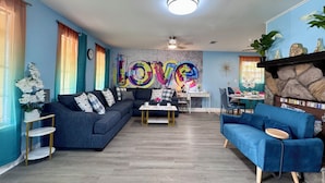 Un salon moderne avec un canapé d’angle, un fauteuil bleu et une table basse. Un mur est décoré d’une fresque colorée représentant le mot « AMOUR ».