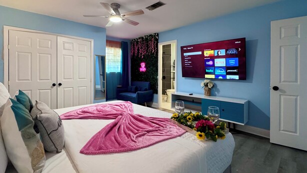 Une chambre d’hôtel avec un lit, un téléviseur diffusant des services de streaming, un ventilateur de plafond et une salle de bain visible à travers une porte ouverte.