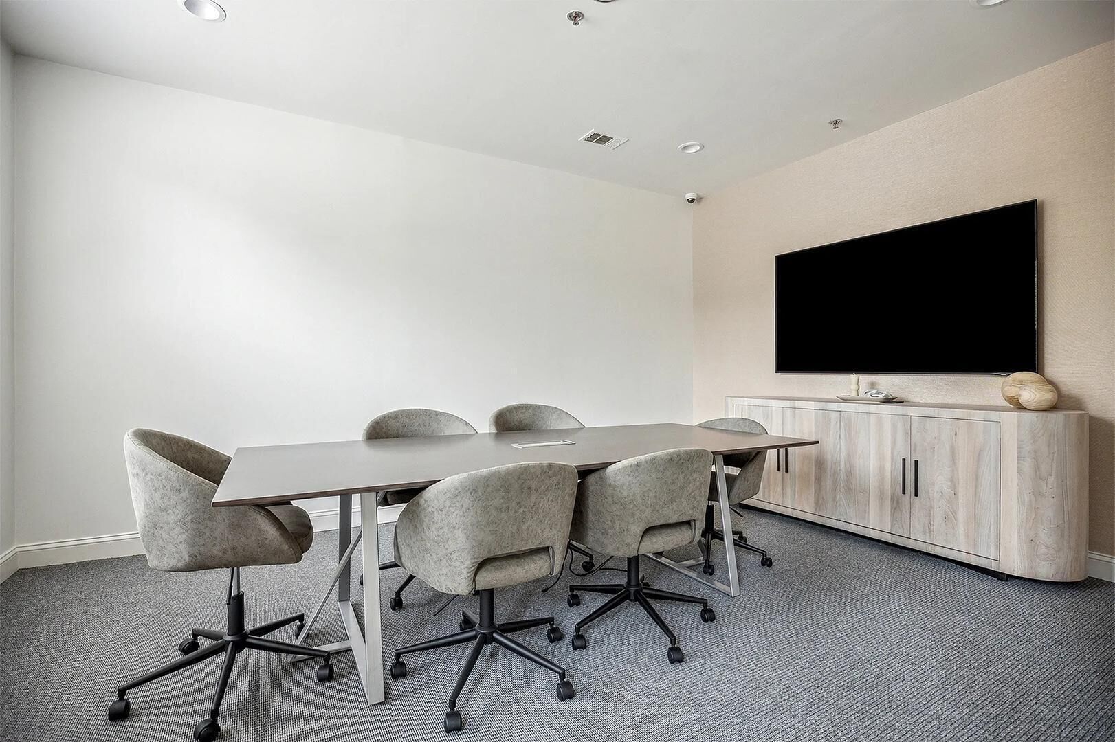 A conference room with a long table, six chairs, a flat-screen TV on the wall, and a wooden cabinet with a decorative item on top.