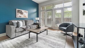 A living room with a grey sofa, a coffee table, and a chair. There is a window with blinds and a painting on the wall. - Landing | Amazing 2BD, Rooftop Lounge, Pool (Cary)