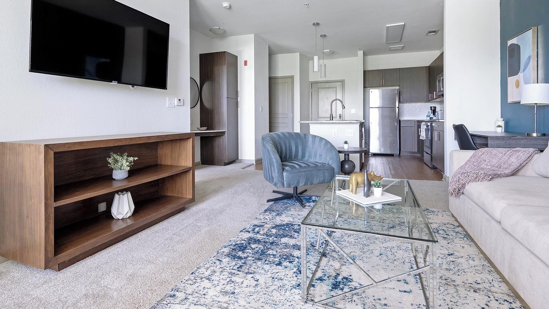 A modern living room with a flat-screen TV, a glass coffee table, a blue patterned rug, and a grey sofa.