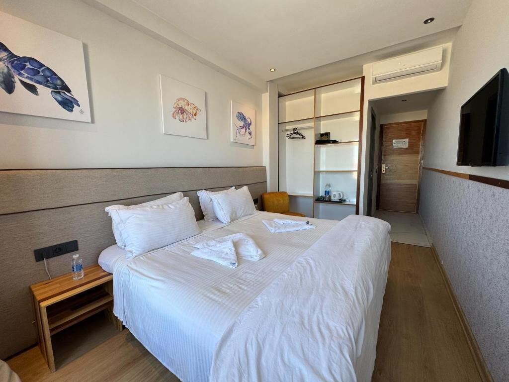 Ein Hotelzimmer mit einem großen Bett, einem Nachttisch mit einer Flasche, einem an der Wand befestigten Fernseher und Bildern an den Wänden.