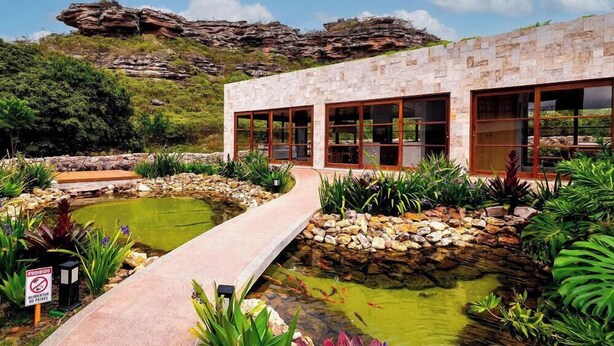 Un edificio moderno con amplios ventanales, fachada de piedra y un jardín ajardinado con un estanque y un sendero.
