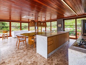 A spacious kitchen with a large island, wooden ceiling, and large windows offering a view of greenery.