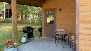 Una cabaña de madera con un porche cubierto, una parrilla de barbacoa y una mesa con sillas.