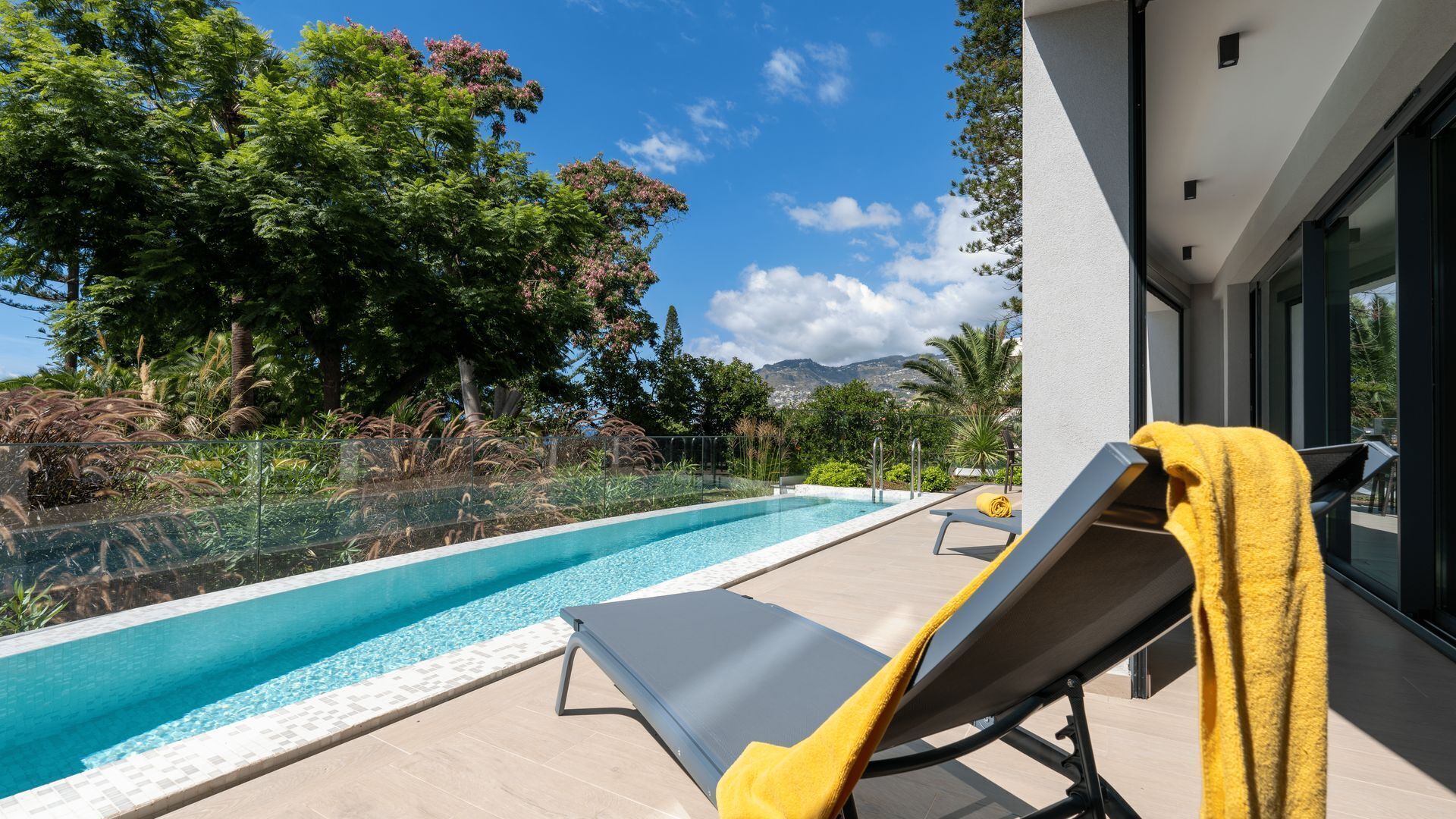 Ein moderner Poolbereich im Freien mit freiem Blick auf Berge im Hintergrund, ein Liegestuhl mit gelbem Handtuch und eine verglaste Terrasse.