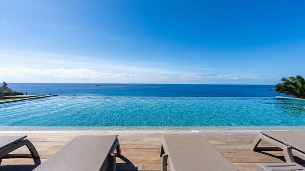 Una piscina infinita con agua azul cristalina, terraza de madera y sillones de descanso.
