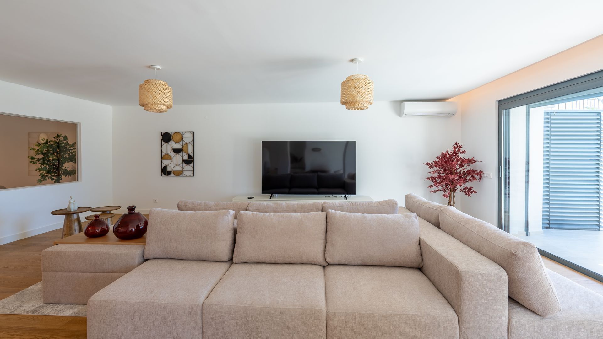 Un moderno salón con un sofá seccional beige, un televisor de pantalla plana, dos lámparas colgantes tejidas y un amplio ventanal con persianas.