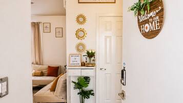 Habitación luminosa con cama, escritorio con planta, espejo en la pared y puerta con un cartel que dice 'Bienvenido a Nuestro Hogar'.