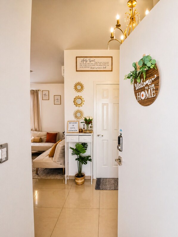 A well-lit room with a bed, a desk with a plant, a wall-mounted mirror, and a door with a 'Welcome to Our Home' sign. - Casa Campos: Perfect Blend of Comfort and Elegance! (San Miguel)