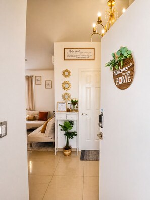 Habitación luminosa con cama, escritorio con planta, espejo en la pared y puerta con un cartel que dice 'Bienvenido a Nuestro Hogar'.