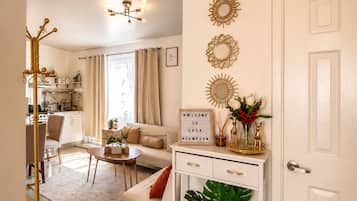 Una sala moderna con un sofá, una mesa de centro y un espejo decorativo. Hay una puerta blanca con un cartel arriba y una planta en el suelo.