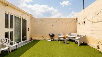 A backyard with artificial grass, white chairs, a table, and string lights.