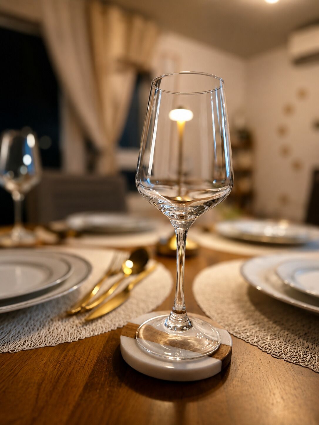 A dining table with a clear glass wine glass on a coaster, surrounded by plates and cutlery.