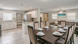 A modern dining area with a large table set for six, surrounded by chairs. There is a kitchen area with white cabinets and a refrigerator. The room has a ceiling fan and a painting on the wall. - Melbourne 6 bed/2 bath Sleeps 12 BEDS 3King/3Qn/2twin (Melbourne)