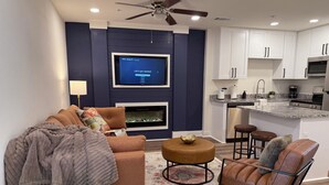 Een moderne woonkamer met een bank, fauteuil, hocker, een open haard en een keuken met witte kasten en een aanrecht.