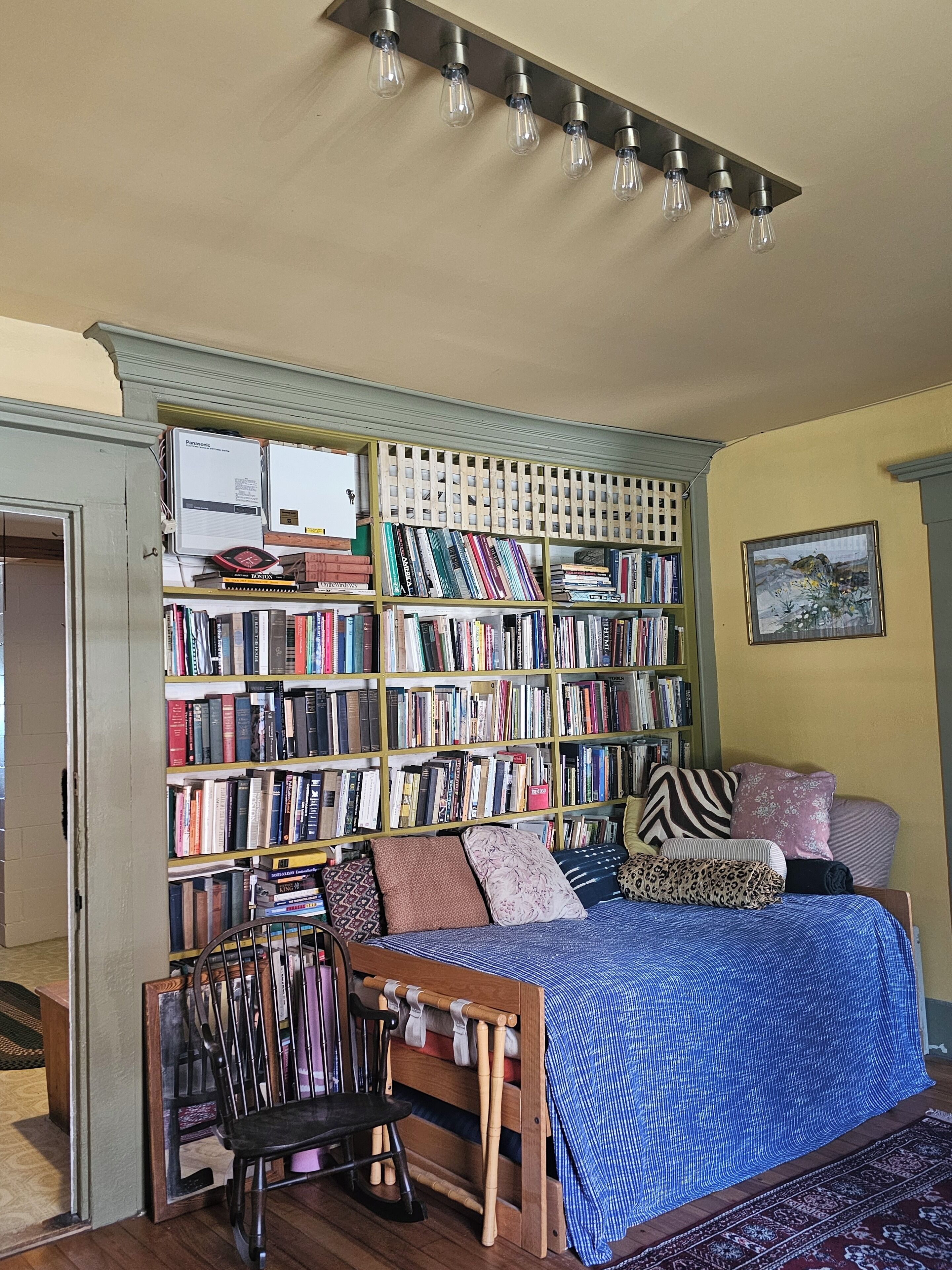 Une pièce chaleureuse avec une grande bibliothèque remplie de livres, une chaise en bois, un lit recouvert d’une couverture bleue et un tableau accroché au mur.