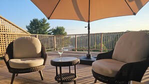 Een terras buiten met rieten stoelen en een tafel, een grote parasol zorgt voor schaduw, en zicht op bomen en een hek op de achtergrond.
