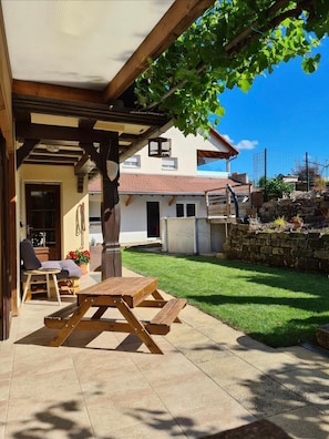 Une terrasse couverte avec une table et des chaises en bois, un mur en pierre et une pelouse verdoyante.