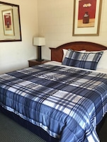 Una cama bien tendida con colcha azul y blanca a cuadros, cabecera de madera y mesita de noche con lámpara.