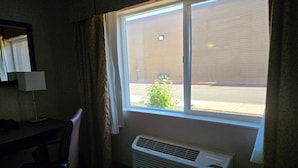 Habitación de hotel con escritorio, silla, unidad de aire acondicionado y ventana con vistas a un edificio y a la calle.