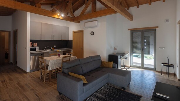 A modern living room with a grey sectional sofa, wooden flooring, and a kitchen area with cabinets and appliances. - Outeiro 7 (Penacova)