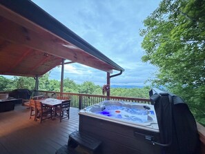 Un bain à remous sur une terrasse en bois, avec une table et des chaises, entouré d’arbres et sous un ciel nuageux.