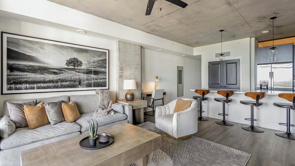 A modern living room with a large black and white landscape painting, a sofa with cushions, a coffee table with plants, a side table with a lamp, and a kitchen area with bar stools and cabinets. - Landing | Amazing 2BD, Gym (St. Petersburg)