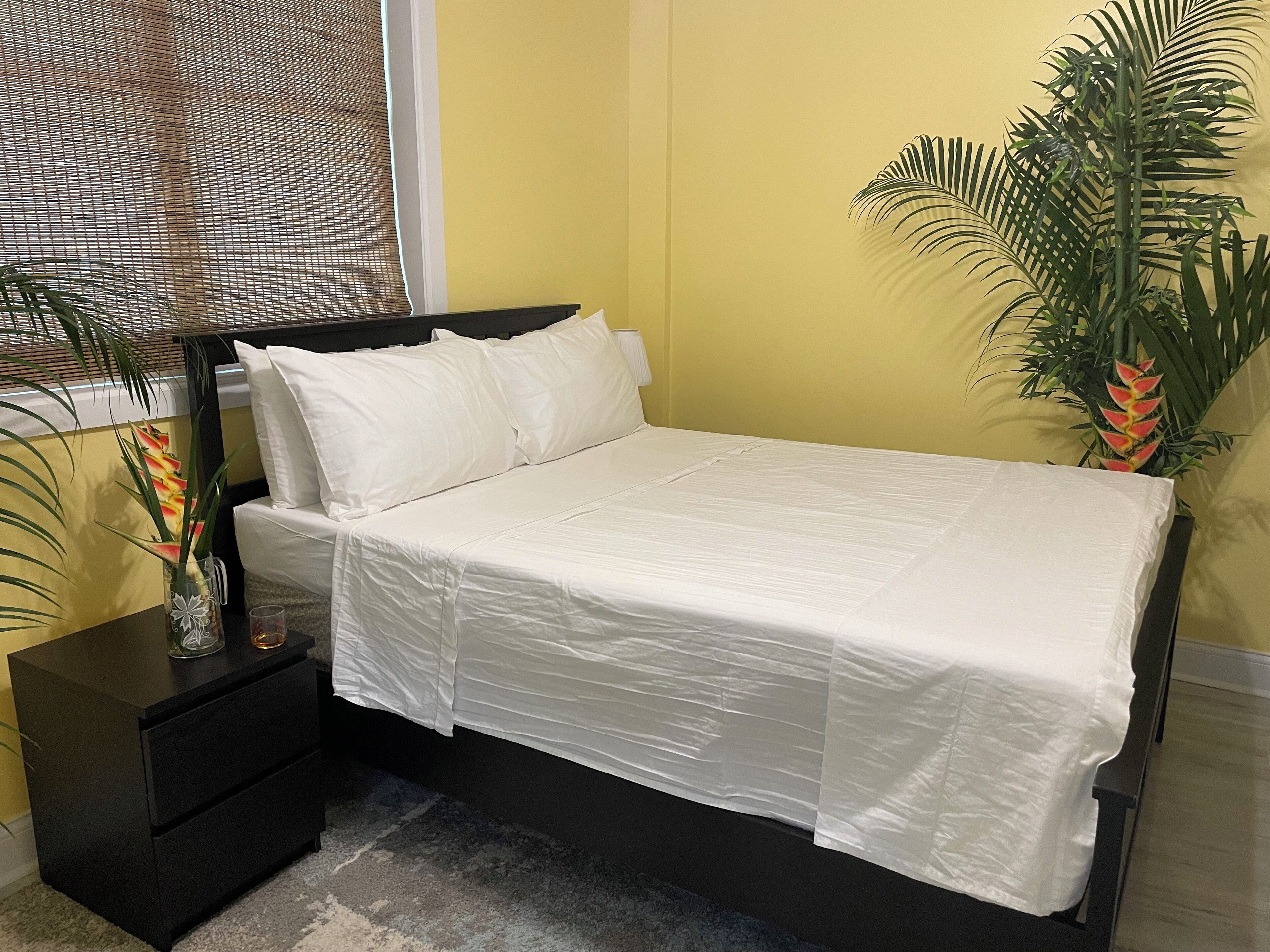 A bed with white bedding, a black nightstand with drawers, and a potted plant beside the bed.