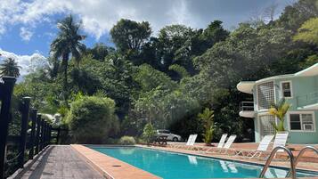 A pool with a wooden deck and white lounge chairs, surrounded by lush greenery and a building with a balcony.