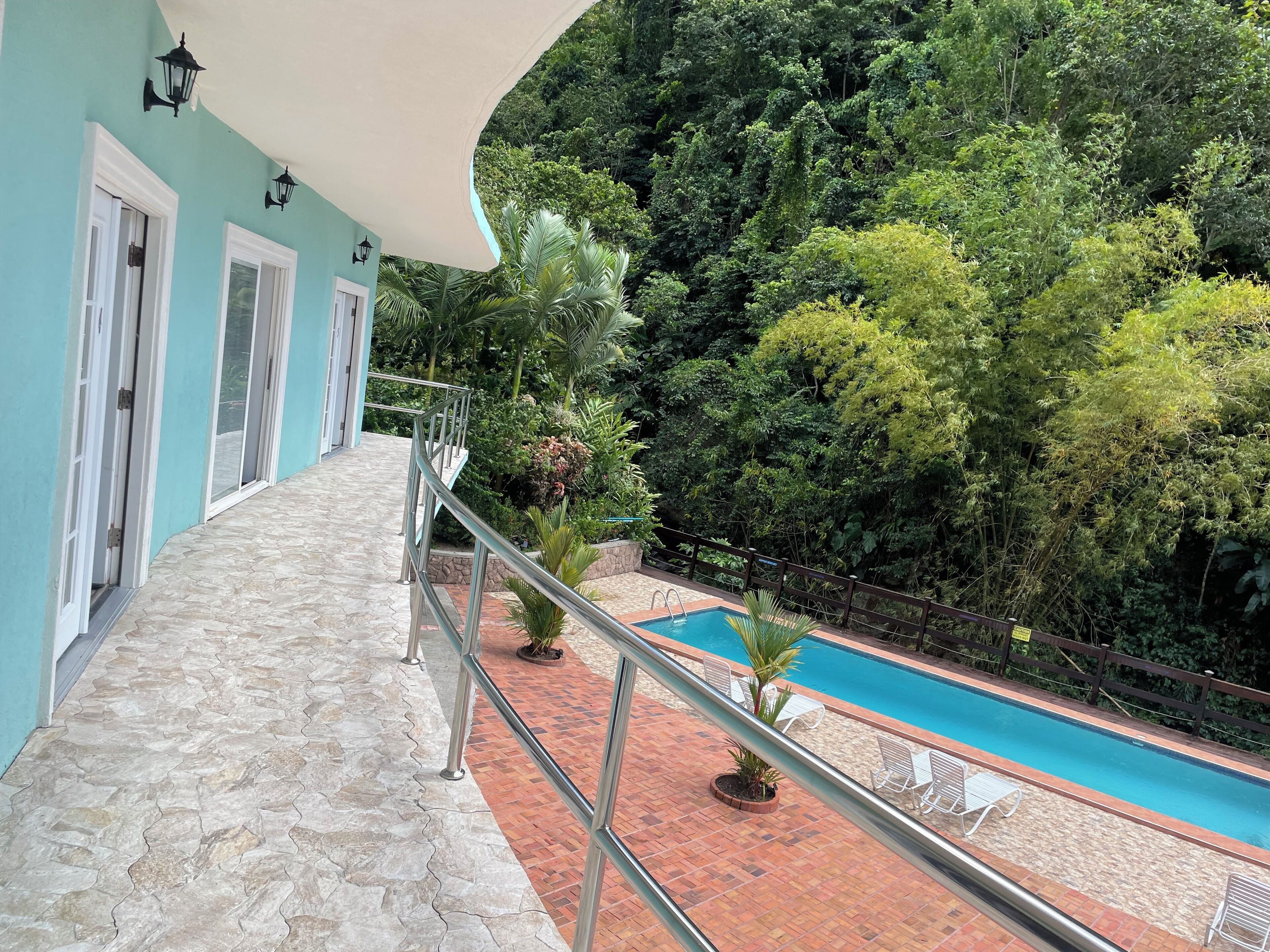 Un'area piscina con un vialetto in pietra che conduce alla piscina, circondata da vegetazione rigogliosa e un edificio dalle pareti color acquamarina e porte bianche.