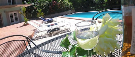 A poolside area with a lounge chair, a glass of drink, and a bottle of wine on a metal table.