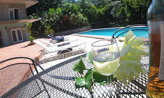 Zona de piscina con una silla de descanso, una bebida en un vaso y una botella de vino sobre una mesa de metal.