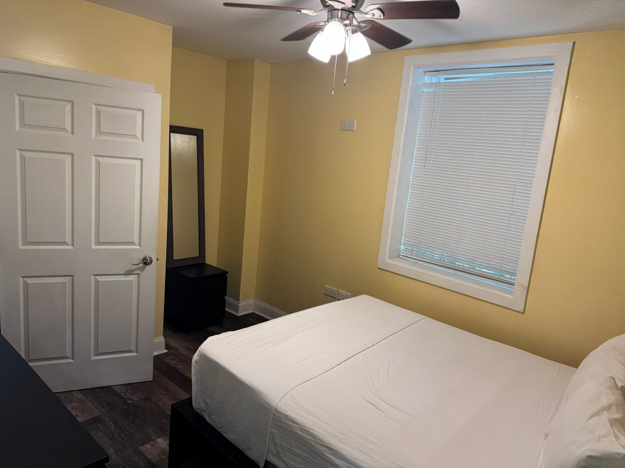 A bedroom with a bed, a ceiling fan, a window with blinds, a door, and a mirror.