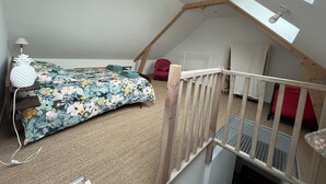 Une chambre avec un plafond en pente, un lit recouvert d’une couvre-lit à motifs floraux, une chaise rouge et une rambarde en bois.