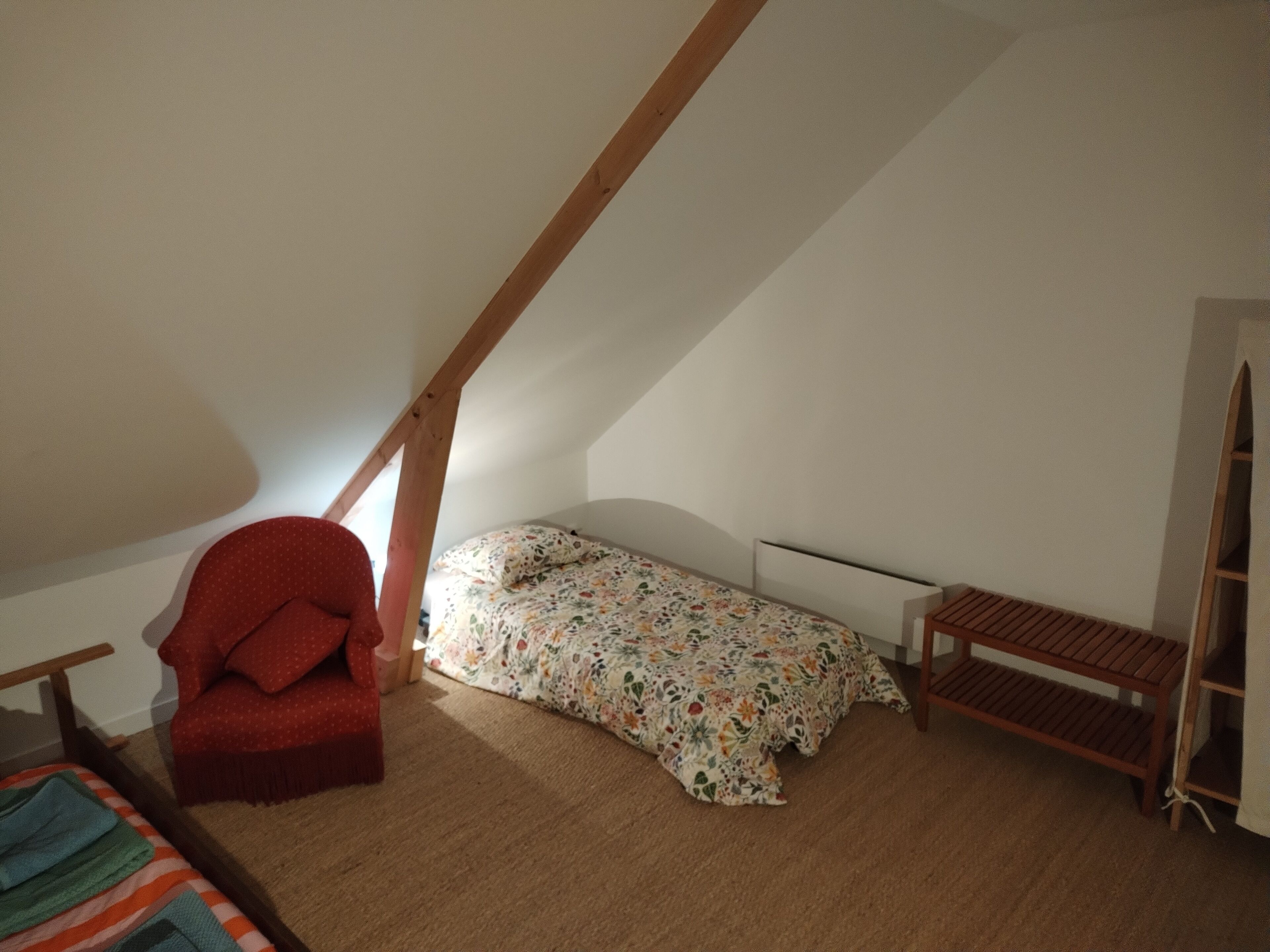 Une petite chambre mansardée, chaleureuse et accueillante, avec un plafond en pente, un lit simple avec une literie à motifs floraux, une chaise rouge et un banc en bois.