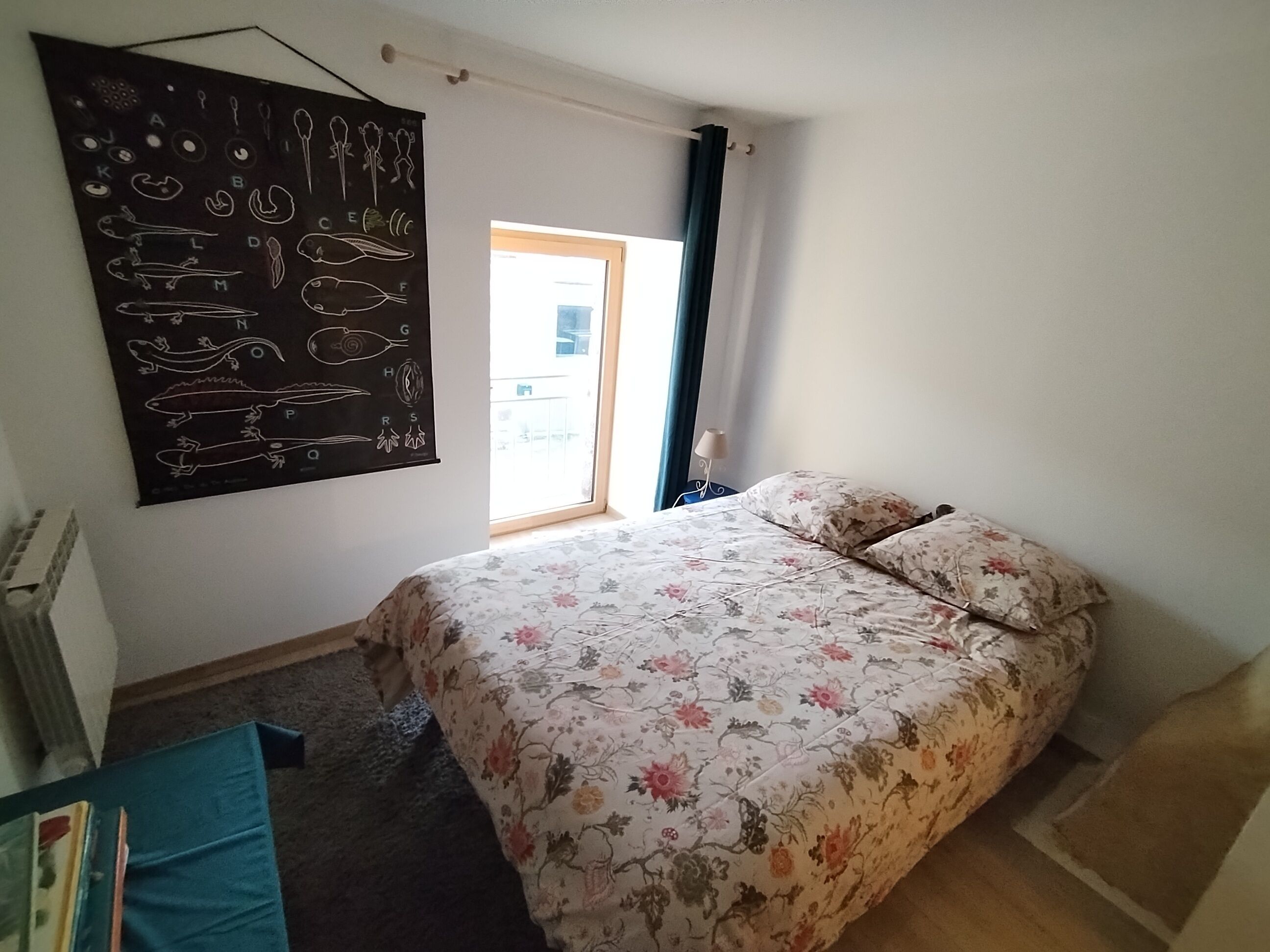 A bedroom with a floral bedspread, a large poster on the wall, and a window with curtains.