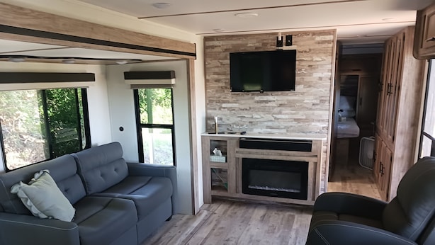 A modern RV interior with a grey sofa, a black leather chair, a flat-screen TV mounted on a stone wall, and a small kitchenette area.